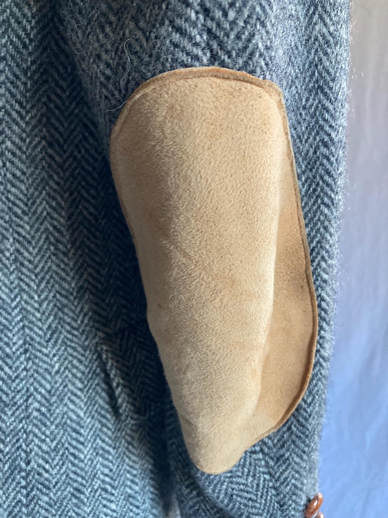 May include: A gray and white herringbone patterned blazer sleeve featuring a large, oval-shaped tan suede elbow patch stitched onto the fabric. The sleeve is textured with a classic woven pattern and includes a visible brown button at the cuff.