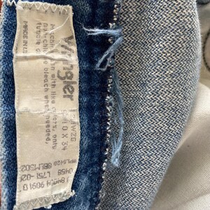 Pu&ograve; includere: Primo piano di una sezione di tessuto denim blu con un'etichetta bianca. L'etichetta recita "Wrangler" e include istruzioni per il lavaggio e informazioni sulle taglie. Il denim ha un aspetto sbiadito.