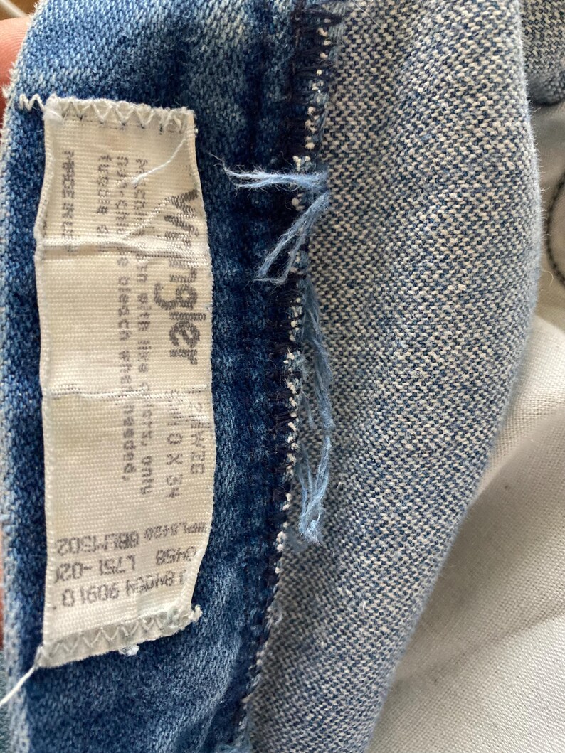 Pu&ograve; includere: Primo piano di una sezione di tessuto denim blu con un'etichetta bianca. L'etichetta recita "Wrangler" e include istruzioni per il lavaggio e informazioni sulle taglie. Il denim ha un aspetto sbiadito.