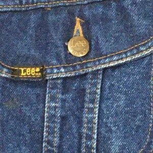 Op de afbeelding: Close-up van een blauw denim jack met een messingknop met het Lee-logo. De knop is bevestigd aan een zakklep.