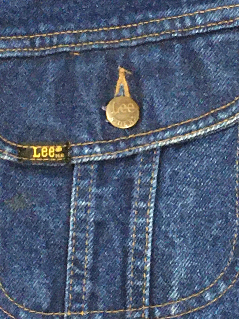 Op de afbeelding: Close-up van een blauw denim jack met een messingknop met het Lee-logo. De knop is bevestigd aan een zakklep.
