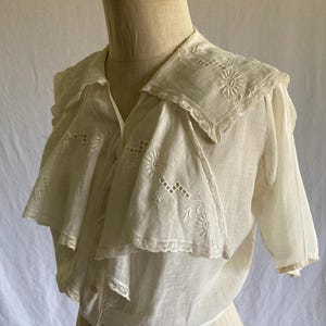May include: A vintage white blouse with a unique layered collar. The collar and upper chest feature delicate embroidered floral patterns and lace trim. The short-sleeved blouse is displayed on a mannequin.