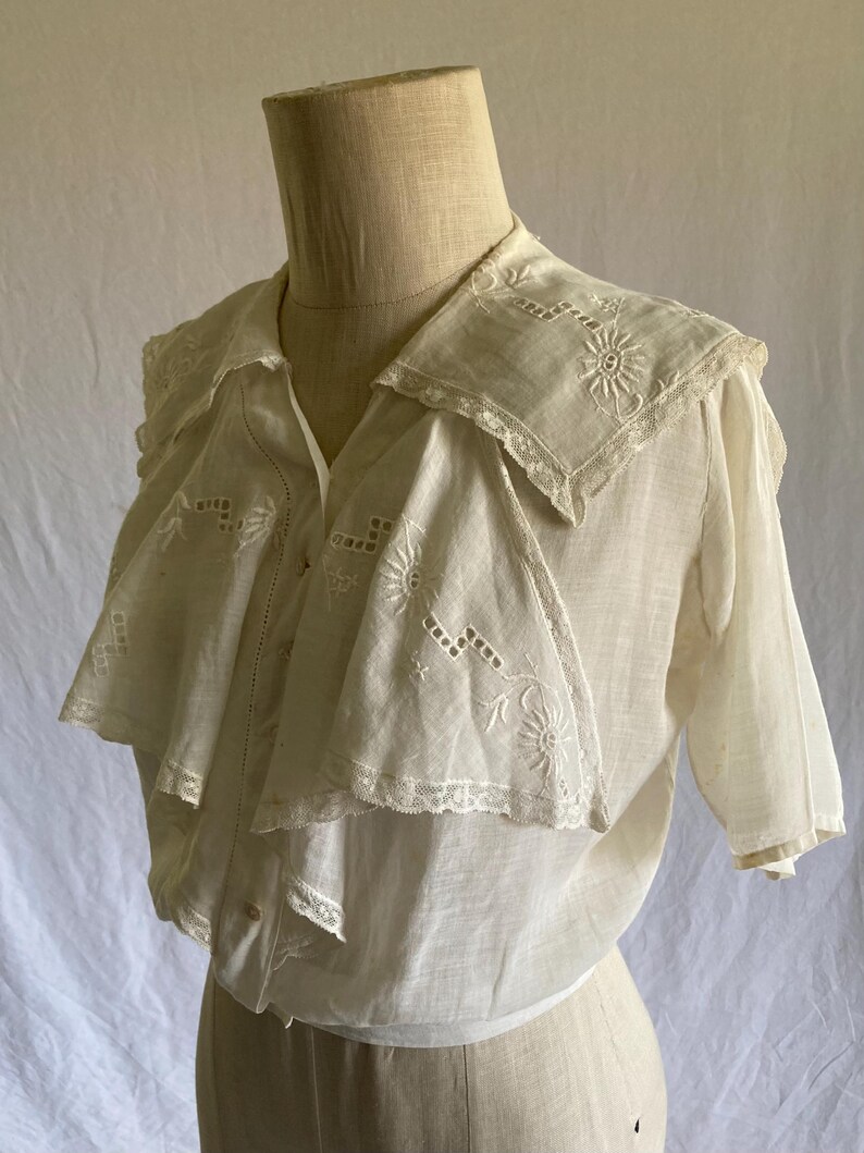 May include: A vintage white blouse with a unique layered collar. The collar and upper chest feature delicate embroidered floral patterns and lace trim. The short-sleeved blouse is displayed on a mannequin.