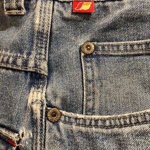 K&ouml;nnte beinhalten: Nahaufnahme einer hellblauen Jeans mit sichtbaren N&auml;hten und Details der Taschen. Mit einem roten Etikett mit einem gelben Blattmuster. Die Jeans hat Metallkn&ouml;pfe und ein klassisches F&uuml;nf-Taschen-Design.