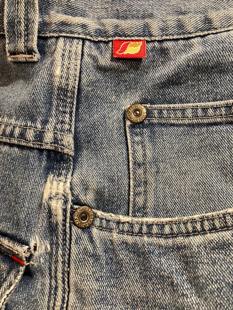 K&ouml;nnte beinhalten: Nahaufnahme einer hellblauen Jeans mit sichtbaren N&auml;hten und Details der Taschen. Mit einem roten Etikett mit einem gelben Blattmuster. Die Jeans hat Metallkn&ouml;pfe und ein klassisches F&uuml;nf-Taschen-Design.