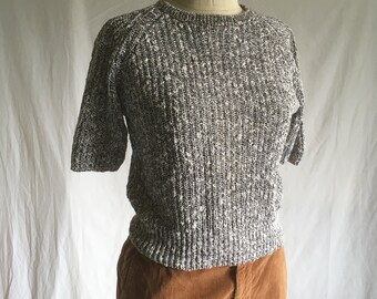May include: A short-sleeved sweater with a speckled gray and white pattern. The sweater is on a mannequin and is paired with brown corduroy pants.