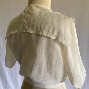 May include: Cream-colored vintage blouse with a wide, lace-trimmed collar. The blouse features short sleeves and a gathered waist. The fabric appears lightweight and semi-sheer, with visible stitching details. The garment is displayed on a mannequin.