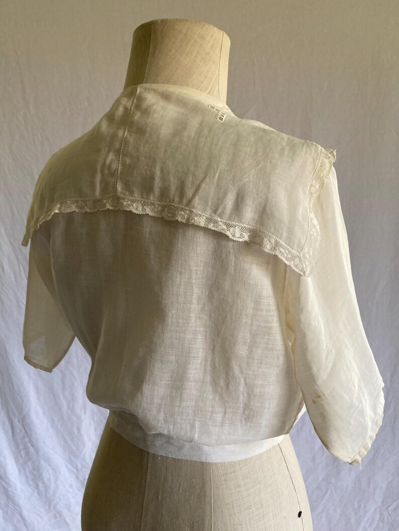 May include: Cream-colored vintage blouse with a wide, lace-trimmed collar. The blouse features short sleeves and a gathered waist. The fabric appears lightweight and semi-sheer, with visible stitching details. The garment is displayed on a mannequin.
