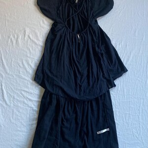 May include: A black dress with a unique design. The dress features a ruffled neckline, short, puffy sleeves, and an open back with crisscross straps. The skirt has a layered, asymmetrical hemline. A silver tag is attached to the skirt.