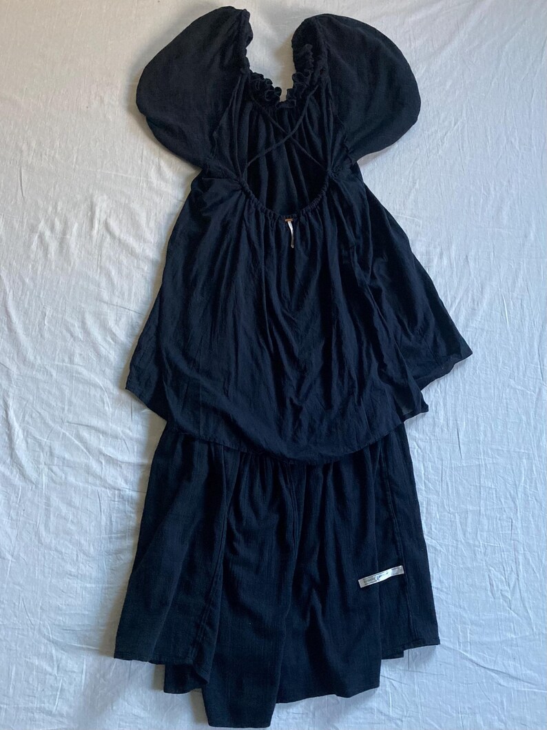 May include: A black dress with a unique design. The dress features a ruffled neckline, short, puffy sleeves, and an open back with crisscross straps. The skirt has a layered, asymmetrical hemline. A silver tag is attached to the skirt.
