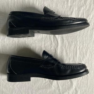 May include: A pair of black leather penny loafers. The shoes feature a classic design with a rounded toe, a strap across the vamp, and a low heel. The shoes are displayed on a white surface.