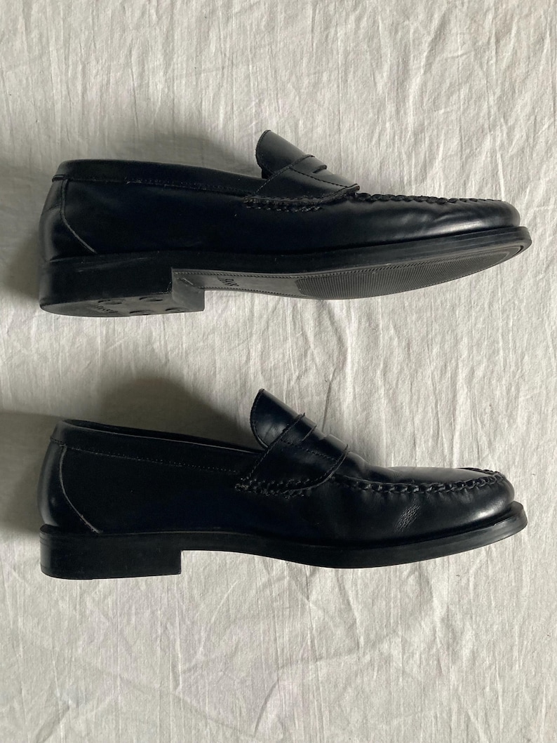 May include: A pair of black leather penny loafers. The shoes feature a classic design with a rounded toe, a strap across the vamp, and a low heel. The shoes are displayed on a white surface.
