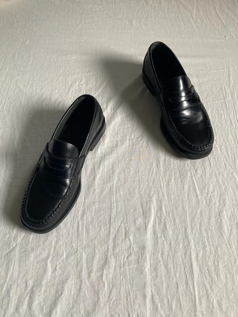 May include: A pair of black leather penny loafers. These classic shoes feature a rounded toe, a strap across the vamp, and a low heel. The leather has a glossy finish. The shoes are displayed on a white surface.