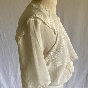 May include: A vintage white blouse with a layered design. The blouse features delicate lace trim along the collar and sleeves. The fabric appears lightweight and semi-sheer, with intricate embroidery details. The blouse is displayed on a mannequin.