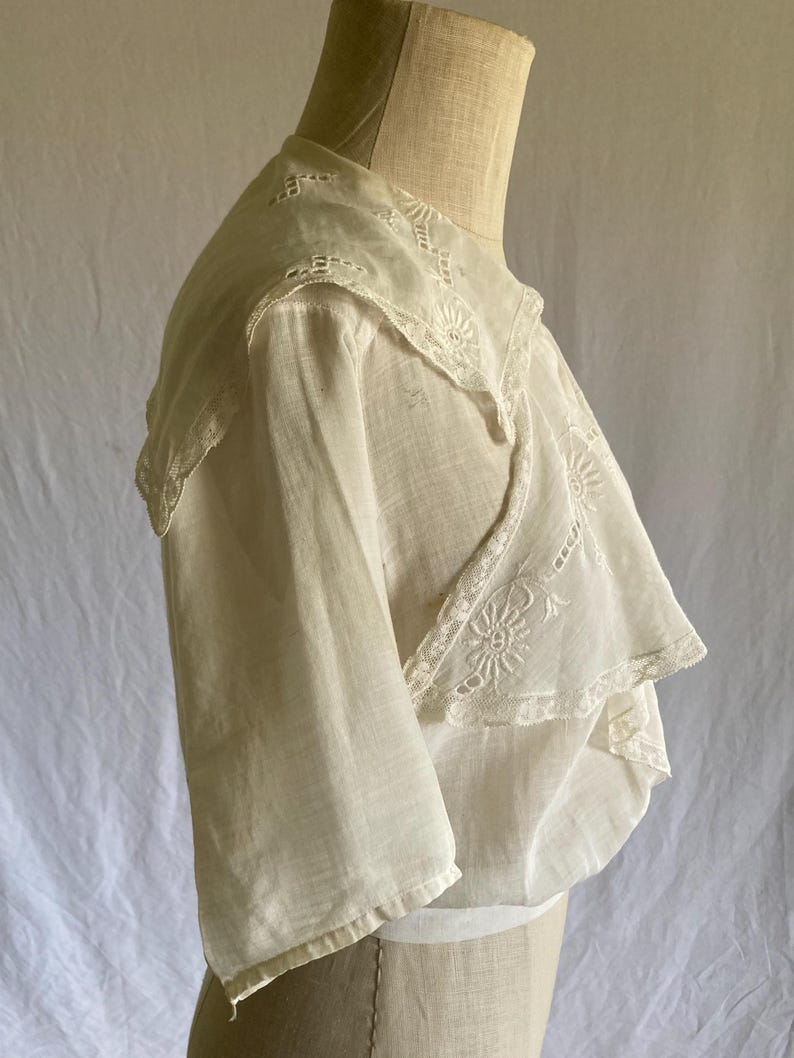May include: A vintage white blouse with a layered design. The blouse features delicate lace trim along the collar and sleeves. The fabric appears lightweight and semi-sheer, with intricate embroidery details. The blouse is displayed on a mannequin.