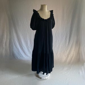 May include: A black dress displayed on a mannequin. The dress features a ruffled neckline, short, puffed sleeves, and a tiered skirt. The fabric appears to be soft and flowing, creating a relaxed silhouette. The dress is a solid black color.