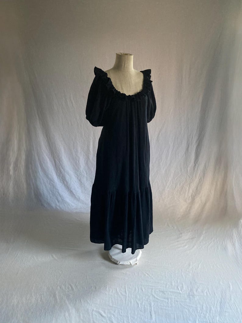 May include: A black dress displayed on a mannequin. The dress features a ruffled neckline, short, puffed sleeves, and a tiered skirt. The fabric appears to be soft and flowing, creating a relaxed silhouette. The dress is a solid black color.
