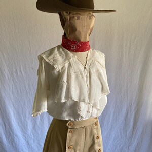 May include: A brown cowboy hat sits atop a paper bag head, with a red bandana around the neck. The outfit includes a white blouse with a ruffled collar and tan high-waisted pants with large buttons. The ensemble is displayed on a mannequin.