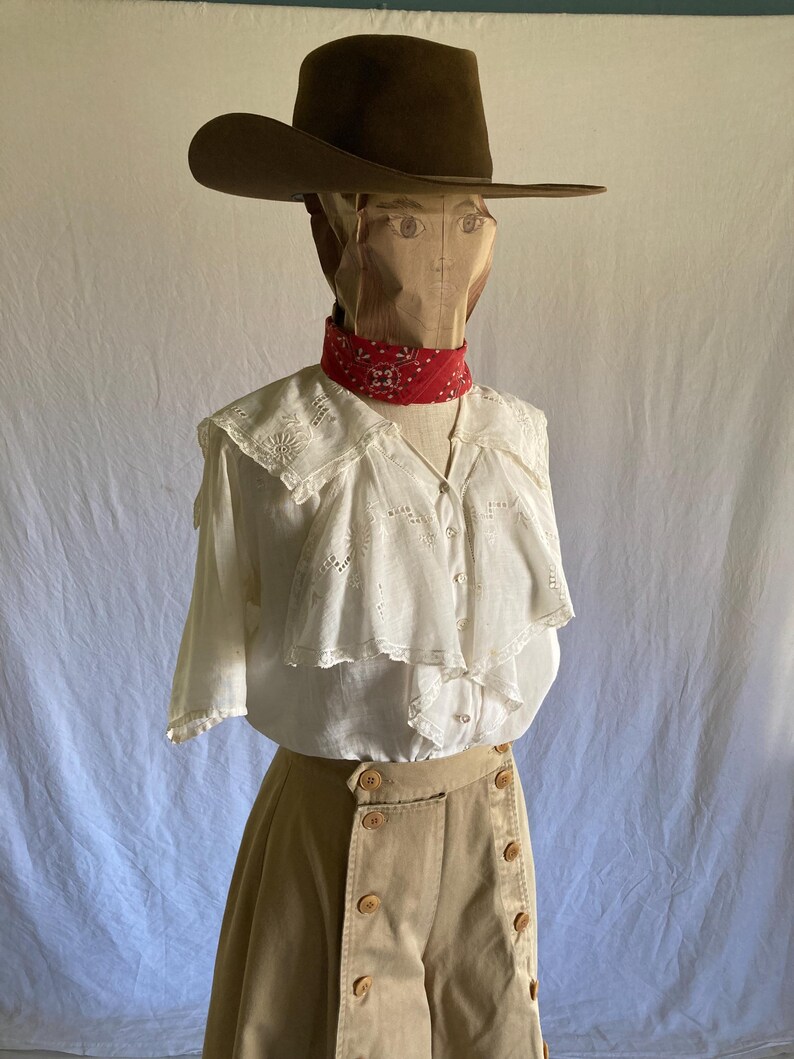 May include: A brown cowboy hat sits atop a paper bag head, with a red bandana around the neck. The outfit includes a white blouse with a ruffled collar and tan high-waisted pants with large buttons. The ensemble is displayed on a mannequin.