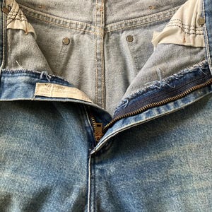 Pu&ograve; includere: Primo piano di jeans di denim blu chiaro con una cerniera in ottone parzialmente aperta. I jeans presentano un classico design a cinque tasche, cuciture visibili e un lavaggio sbiadito. L'etichetta Wrangler &egrave; visibile.