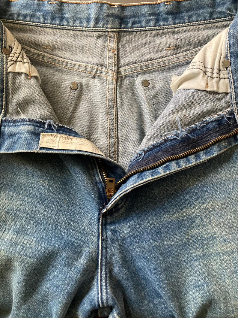 Pu&ograve; includere: Primo piano di jeans di denim blu chiaro con una cerniera in ottone parzialmente aperta. I jeans presentano un classico design a cinque tasche, cuciture visibili e un lavaggio sbiadito. L'etichetta Wrangler &egrave; visibile.