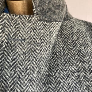 May include: A gray and white herringbone patterned jacket featuring a solid dark gray felted wool collar. The garment is displayed on a mannequin, showcasing the textured weave of the fabric and the structured stitching along the collar edge.