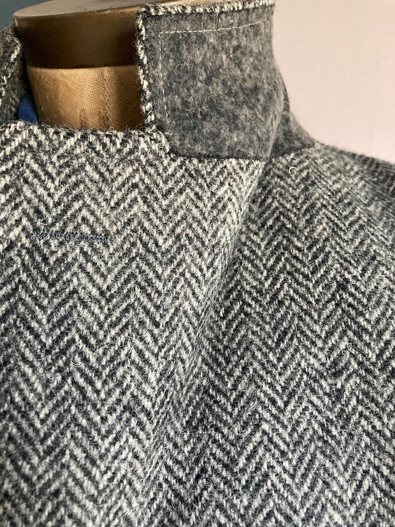 May include: A gray and white herringbone patterned jacket featuring a solid dark gray felted wool collar. The garment is displayed on a mannequin, showcasing the textured weave of the fabric and the structured stitching along the collar edge.