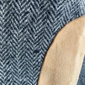 May include: A close-up view of a garment featuring a gray and white herringbone wool fabric pattern. A smooth, tan-colored suede or leather patch is stitched onto the side of the textured, woven material, creating a distinct contrast in both color and surface texture.