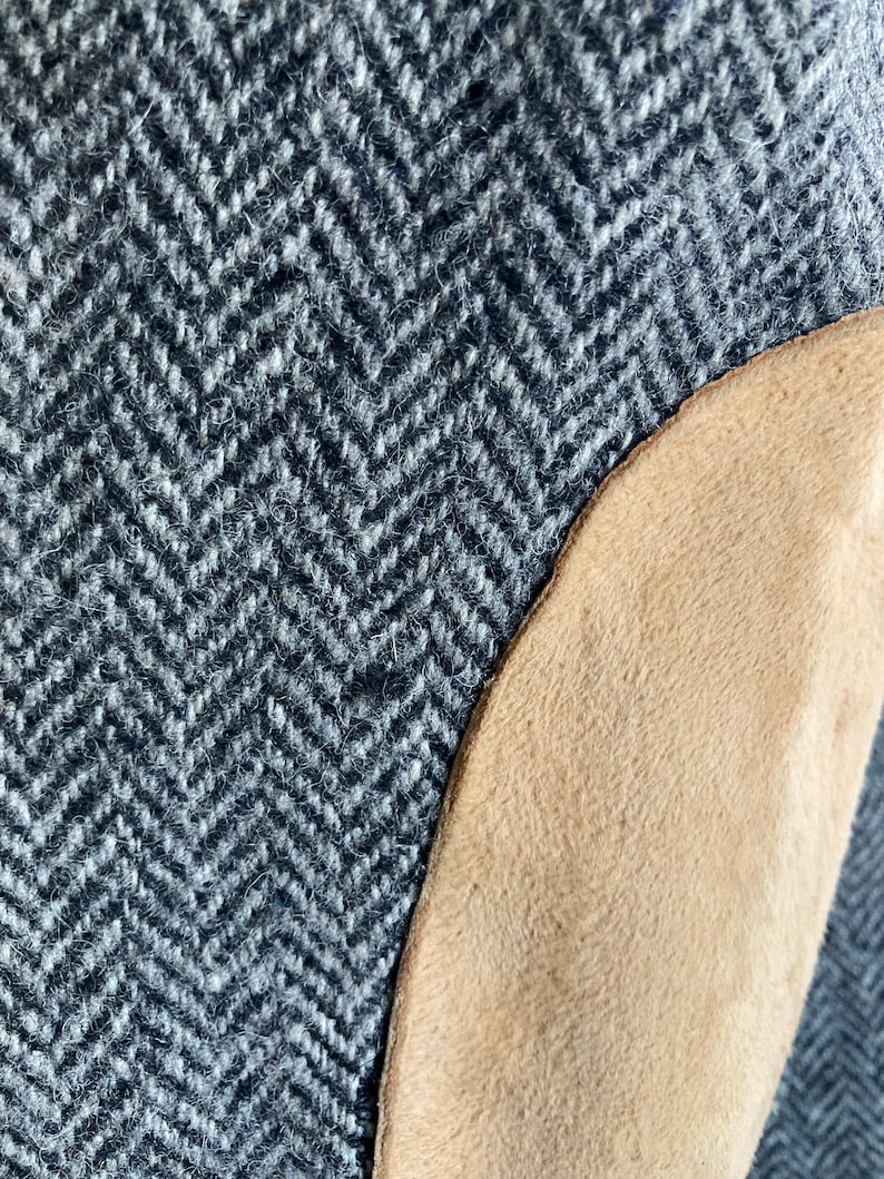 May include: A close-up view of a garment featuring a gray and white herringbone wool fabric pattern. A smooth, tan-colored suede or leather patch is stitched onto the side of the textured, woven material, creating a distinct contrast in both color and surface texture.
