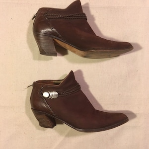 May include: A pair of brown leather ankle boots with a braided strap and a silver buckle. The boots have a block heel and a pointed toe.