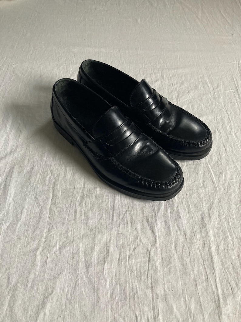 May include: A pair of black leather penny loafers. The shoes feature a classic design with a rounded toe, a strap across the vamp, and a low heel. The leather has a glossy finish.