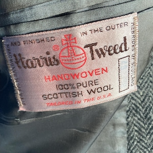 May include: A rectangular fabric label sewn into the interior of a grey herringbone garment. The label reads Harris Tweed, Handwoven, 100% Pure Scottish Wool, and Tailored in the U.S.A., featuring a red orb logo and text in black and red ink.