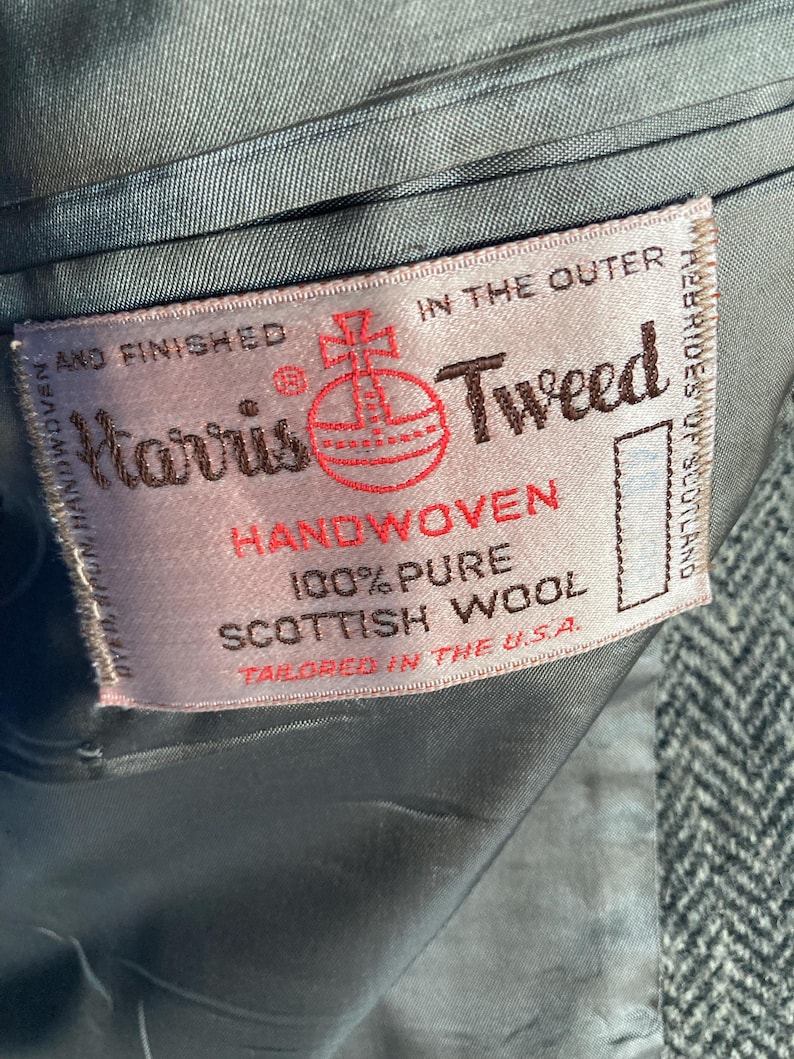 May include: A rectangular fabric label sewn into the interior of a grey herringbone garment. The label reads Harris Tweed, Handwoven, 100% Pure Scottish Wool, and Tailored in the U.S.A., featuring a red orb logo and text in black and red ink.