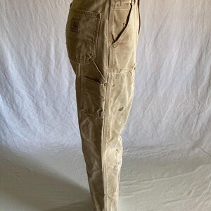 May include: Tan work pants with multiple pockets and a red logo patch. The pants have a worn appearance with visible stains and wear, suggesting they are well-used. The pants are displayed on a mannequin.
