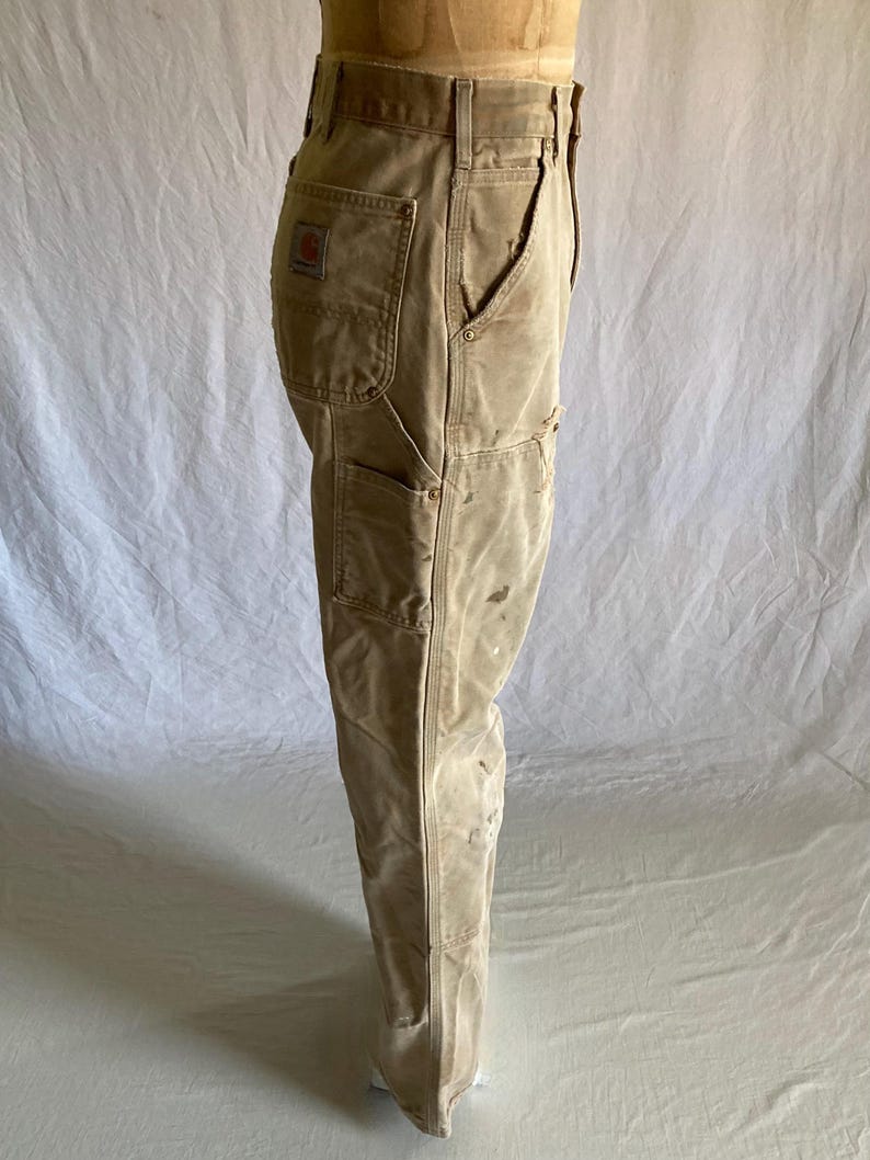 May include: Tan work pants with multiple pockets and a red logo patch. The pants have a worn appearance with visible stains and wear, suggesting they are well-used. The pants are displayed on a mannequin.