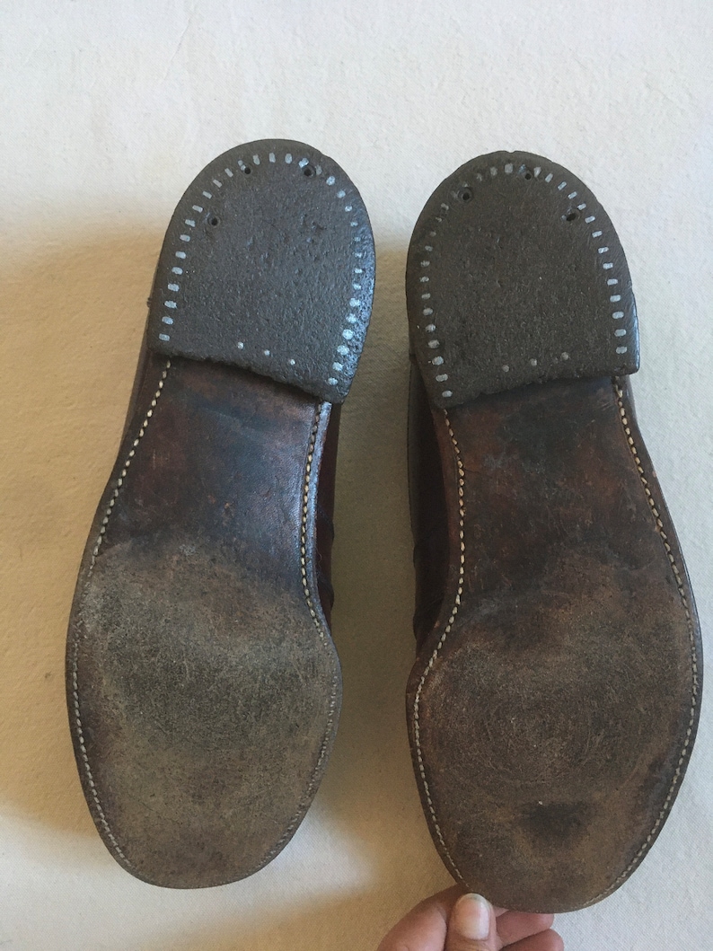 May include: A pair of brown leather shoes with white stitching around the edges of the soles. The soles are worn and have a pattern of small holes.