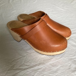 May include: A pair of brown leather clogs with wooden soles and heels. The clogs feature a classic design with a strap across the top and are stitched together with white thread. The shoes are set against a white background.