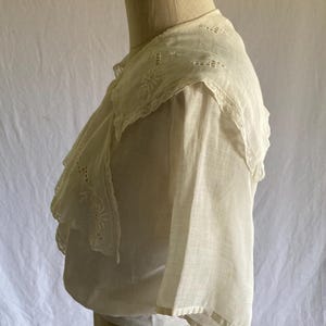 May include: Off-white vintage blouse with a detailed, embroidered collar and ruffled trim. The blouse features delicate floral embroidery and a layered design, showcasing a classic, romantic aesthetic. The sleeves are short and wide.