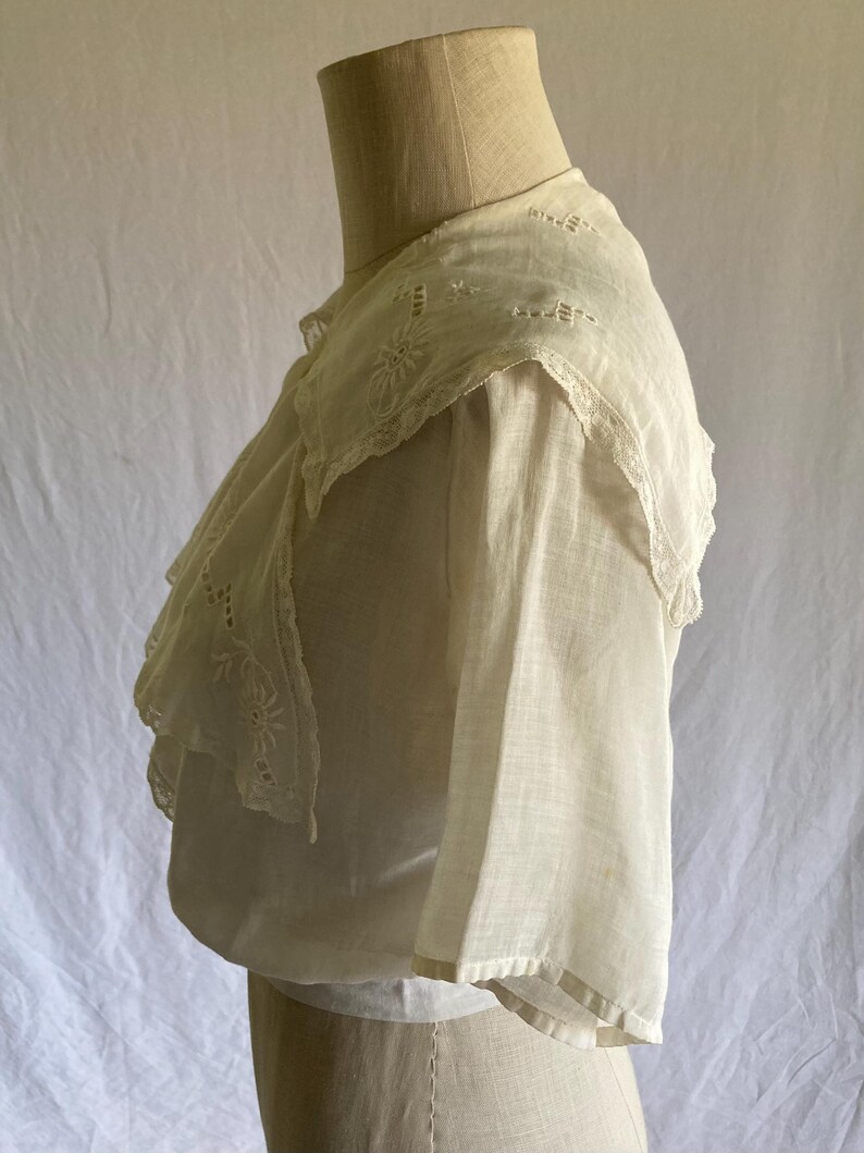 May include: Off-white vintage blouse with a detailed, embroidered collar and ruffled trim. The blouse features delicate floral embroidery and a layered design, showcasing a classic, romantic aesthetic. The sleeves are short and wide.