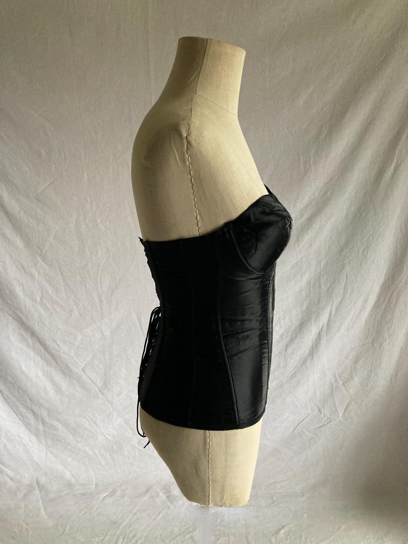May include: A black satin corset with a lace-up back and a boned structure. The corset is displayed on a white mannequin.