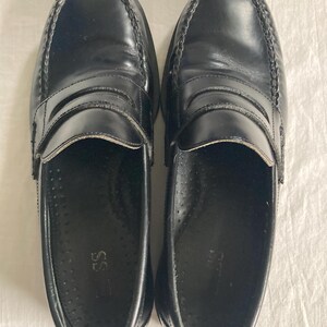 May include: A pair of black leather penny loafers. The shoes have a glossy finish, a stitched vamp, and a strap across the top. The soles are dark and the interior is black. The shoes are displayed on a white surface.