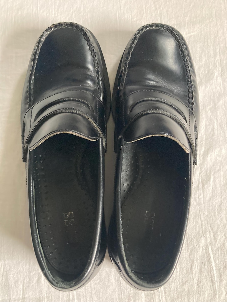 May include: A pair of black leather penny loafers. The shoes have a glossy finish, a stitched vamp, and a strap across the top. The soles are dark and the interior is black. The shoes are displayed on a white surface.