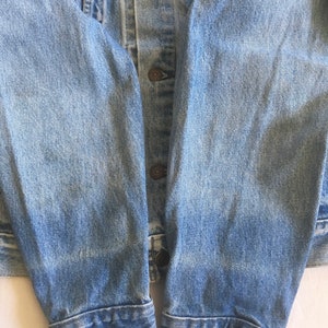 May include: Close-up of the sleeves of a light blue denim jacket. The sleeves are folded up at the cuffs, showing the raw edge of the denim.