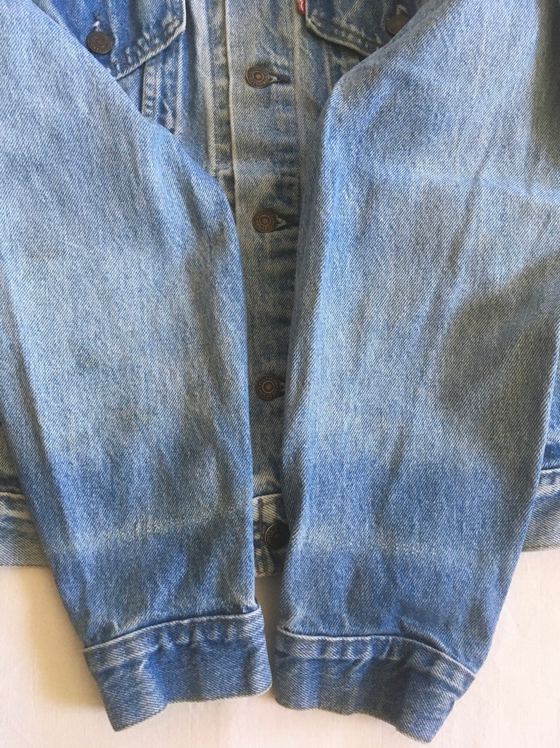 May include: Close-up of the sleeves of a light blue denim jacket. The sleeves are folded up at the cuffs, showing the raw edge of the denim.
