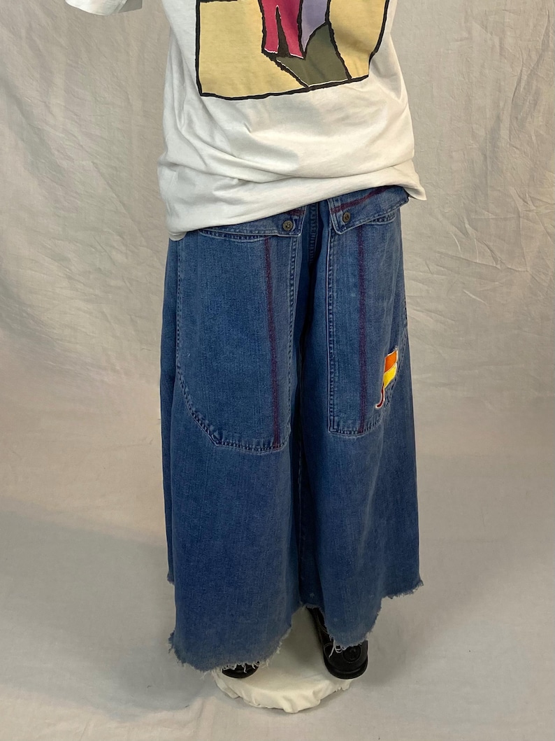 K&ouml;nnte beinhalten: Blauer Jeansrock mit rohem Saum und gro&szlig;en Taschen. Der Rock hat vertikale lila Streifen und einen kleinen Aufn&auml;her mit einem gelb-roten Design. Das Model tr&auml;gt ein wei&szlig;es T-Shirt mit einer bunten Grafik.