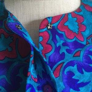 Op de afbeelding: Een blauwe en roze gebloemde zijden blouse met een knoopsluiting en een kraag. De blouse heeft een uniek ontwerp met een bloemenpatroon.