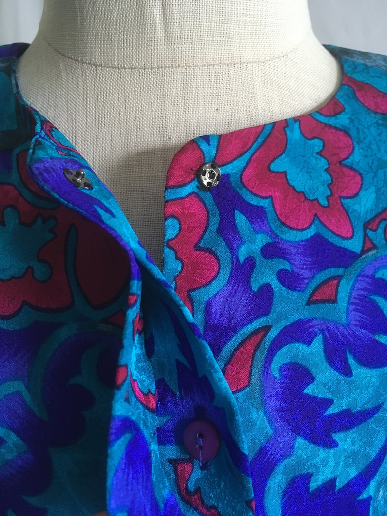 Op de afbeelding: Een blauwe en roze gebloemde zijden blouse met een knoopsluiting en een kraag. De blouse heeft een uniek ontwerp met een bloemenpatroon.