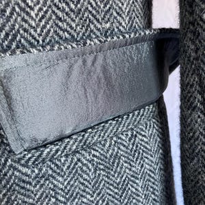 May include: A charcoal gray and white herringbone patterned wool coat featuring a smooth, solid dark gray fabric belt loop detail integrated into the waistline. The textured fabric shows a classic woven chevron design throughout the garment.