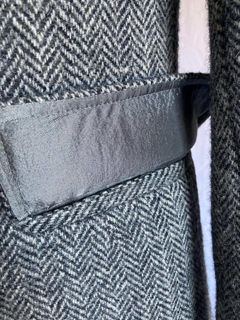 May include: A charcoal gray and white herringbone patterned wool coat featuring a smooth, solid dark gray fabric belt loop detail integrated into the waistline. The textured fabric shows a classic woven chevron design throughout the garment.