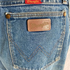 Pu&ograve; includere: Primo piano di jeans blu con una tasca posteriore con una toppa in pelle marrone e il nome del marchio "Wrangler" in rilievo. I jeans hanno un'etichetta rossa con il logo "Wrangler" e rivetti in ottone.